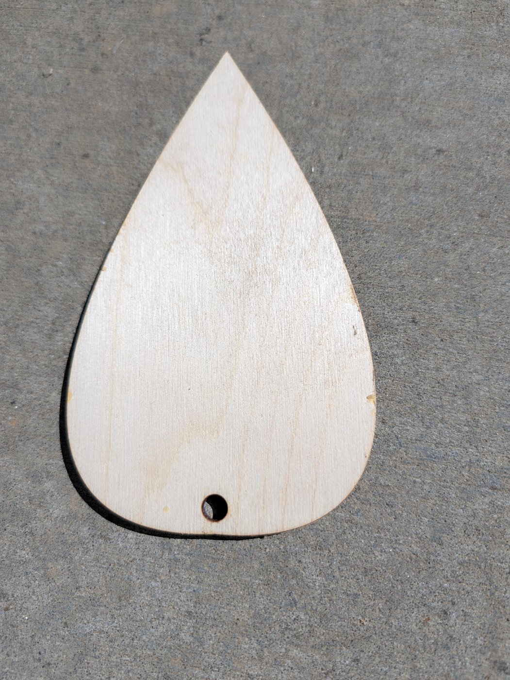 The convex tapered bullroarer blade as built, two laminations glued face to face, lying on concrete: a pale birch teardrop pointed at the top and round at the bottom, with the cord hole near the round end
