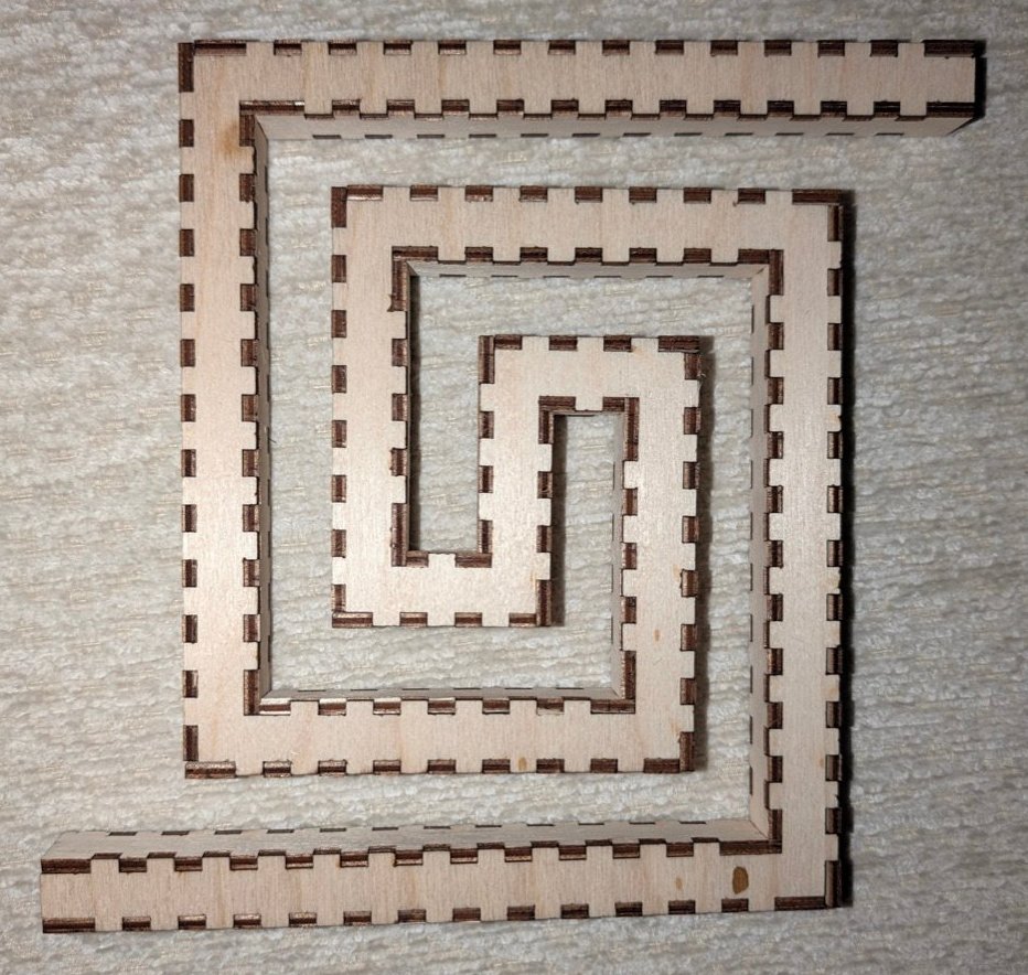 The greek spiral cut in birch ply and press-fit, lying flat: a square-section bore with its finger joints showing dark along every edge, winding in from the top right to a short bar at the centre and back out to the bottom left