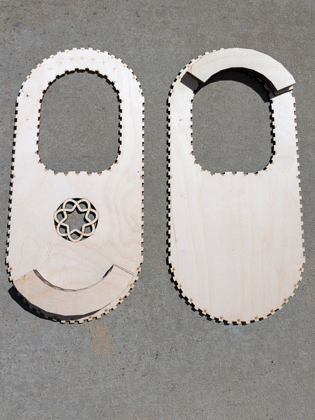 The lyre-harp parts as cut, lying side by side on concrete: on the left the front cheek, a long stadium of birch with finger-jointed edges, the string hole near the top, a row of engraved crosses over it and the seven-fold knot rosette below it, with the curved laminations of the hitch-pin block stacked across its lower end; on the right the plain back cheek, with the laminations of the tuning-pin block stacked over the top of its string hole