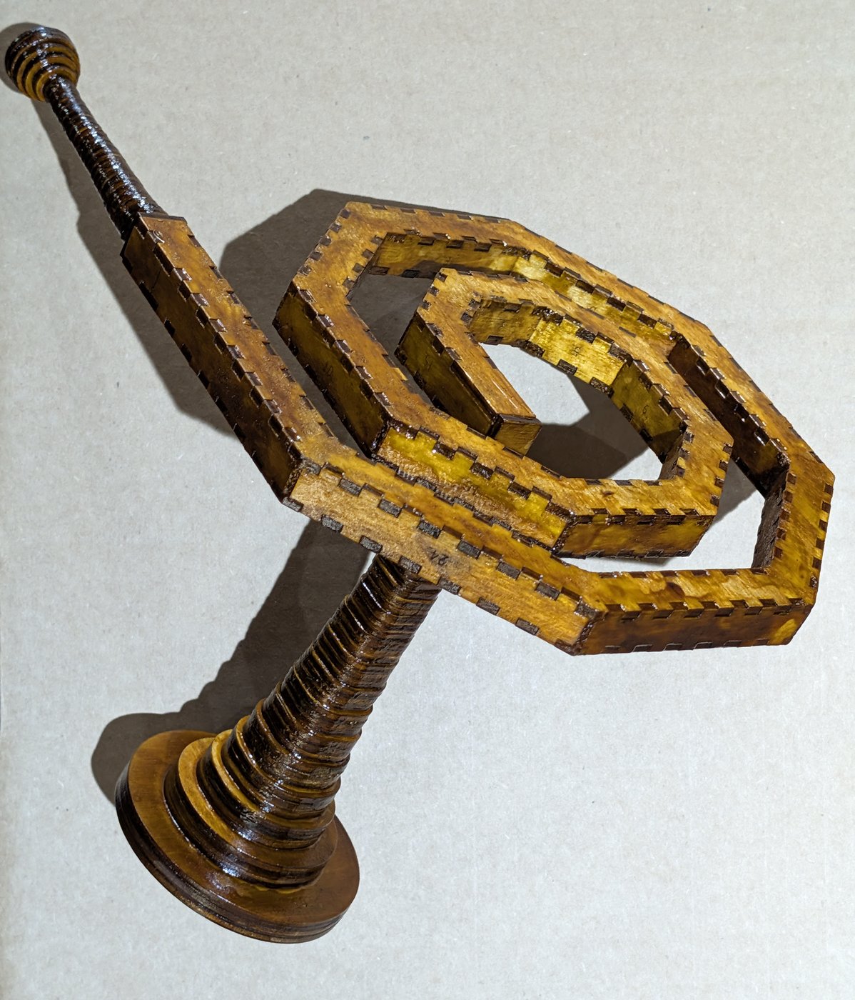 The finished spiral standing upright on its bell on grey card: the bell's round rim flat on the card and its ribbed throat rising into the port at the inner end of the coil, the faceted band winding a little over two turns above it in glossy amber-brown with darker tabs along both rims, and the straight tail leaving the outer end to carry a dark ring-stacked mouthpiece up to the top left