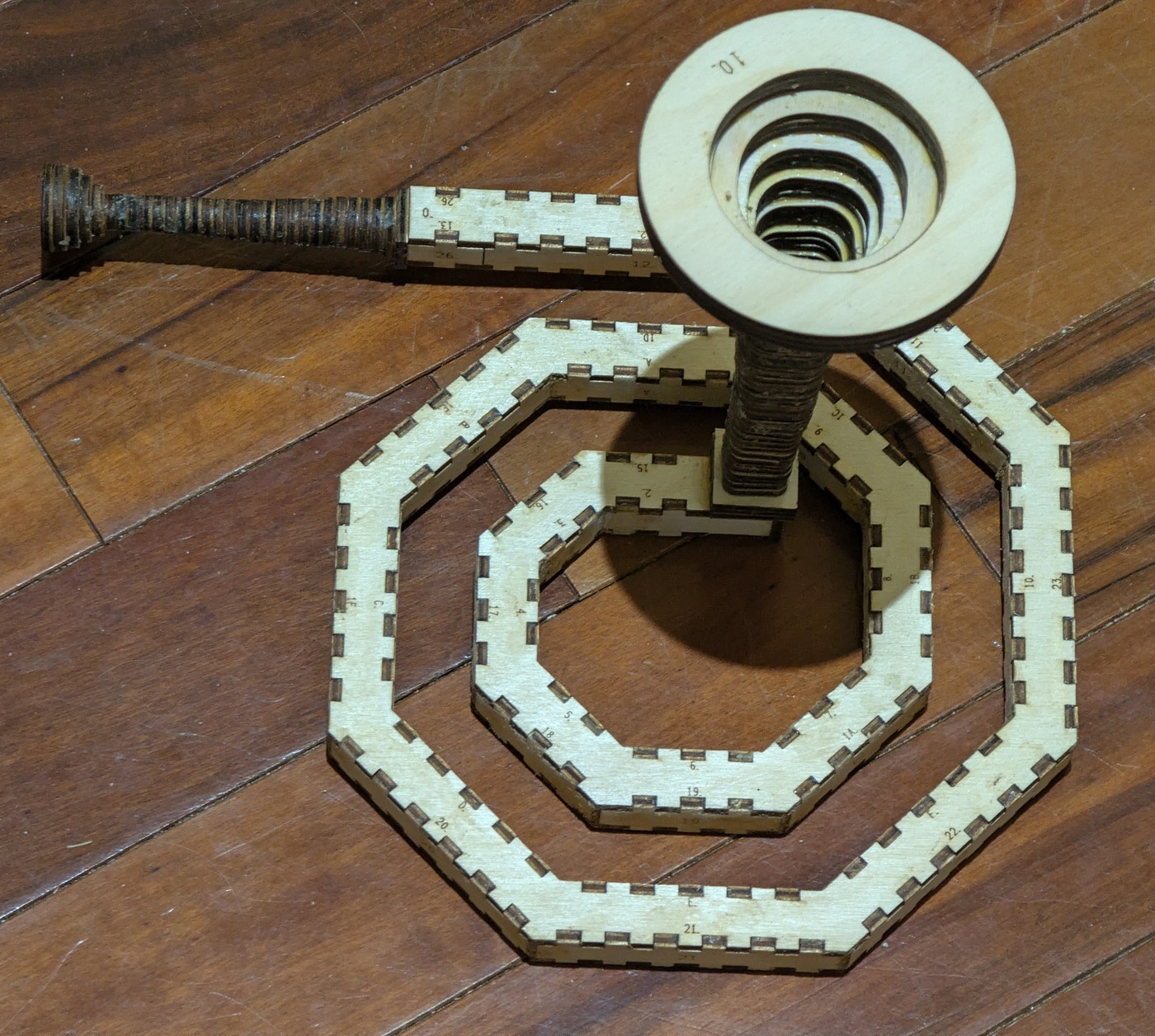 The spiral press-fit on a dark wood floor: a pale ply band winding a little over two turns, cornered at every facet, both rims crenellated with tabs and a number engraved beside each one; a straight tail leaves the outer end and carries a dark, glossy mouthpiece, and a bell engraved 10. stands on a dark ribbed throat at the inner end