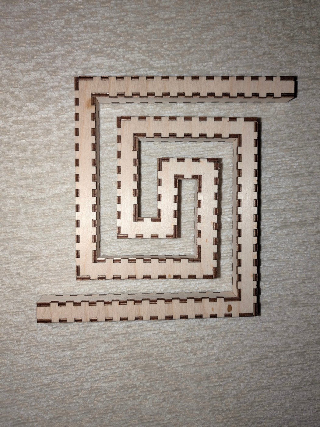 The greek spiral cut in birch ply and press-fit, lying flat: a square-section bore with its finger joints showing dark along every edge, winding in from the top right to a short bar at the centre and back out to the bottom left