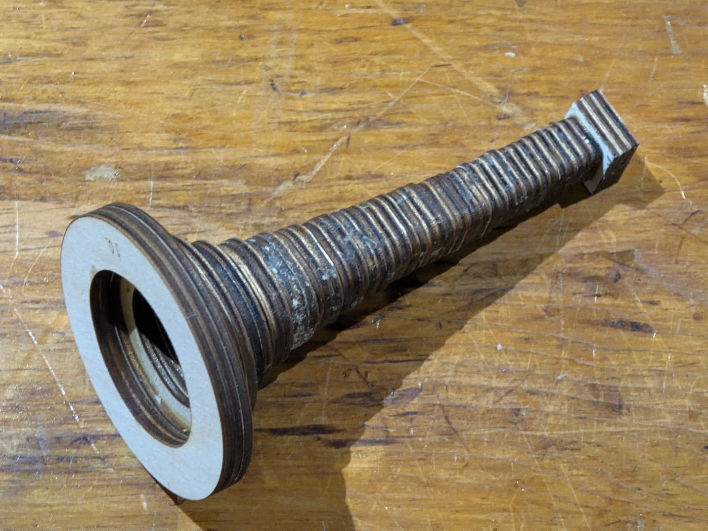 The bell as built, lying on a scratched workbench: a stack of laminated ply rings rising from a small square block at the throat, widening and rounding as it climbs to a flat round rim, every glue line showing as a stripe up the side