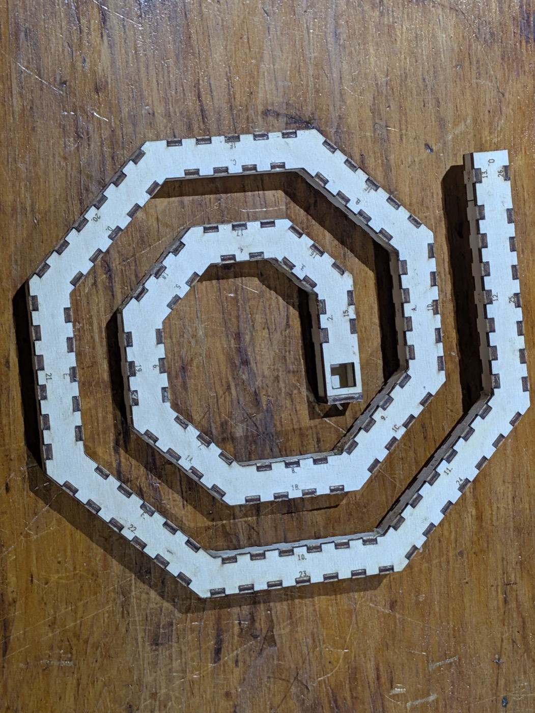 The two cheek plates as cut, one lying on the other on a scratched workbench: a faceted band of pale masked ply winding a little over two turns out from a small centre to a straight tail, both rims crenellated with open notches, a number engraved beside each notch, and a small square hole at the inner end