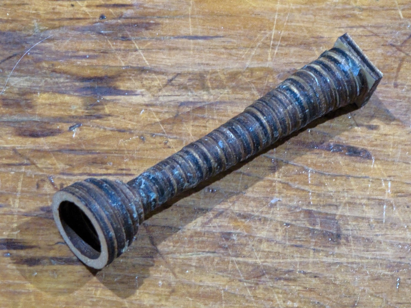 The mouthpiece as built, lying on a scratched workbench: a square block at one end, a long ring-stacked shaft narrowing to a waist, and a small cup opening at the other end, the ply laminations visible as rings along its whole length